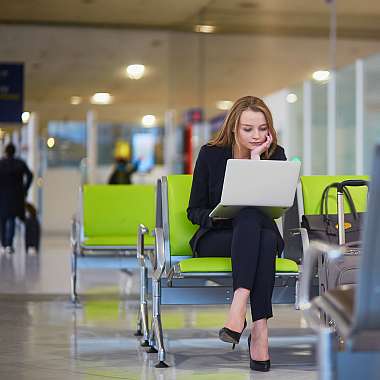 EIne junge Frau im schwarzen Business Dress sitzt an Flughafen, hat ihr Laptop auf den Knien und hat ihre Reise mit einer Reiseversicherung Geschäftsreisen und Versicherungsrechner abgesichert.