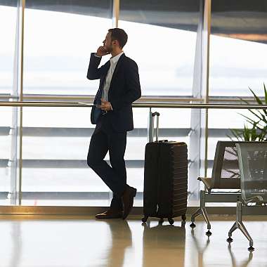 Ein Business Mann im schwarzen Anzug steht in der Wartehalle am Flughafen und hat seine Reise mit einer Reiseversicherung Geschäftsreisen und Versicherungsrechner abgesichert.